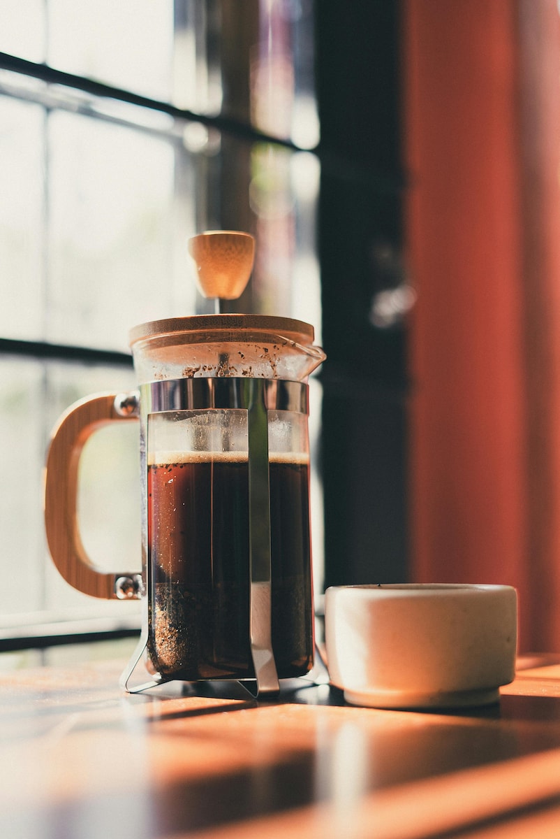 A glass French press of dark coffee on a wooden table, lit by warm morning light through a window.